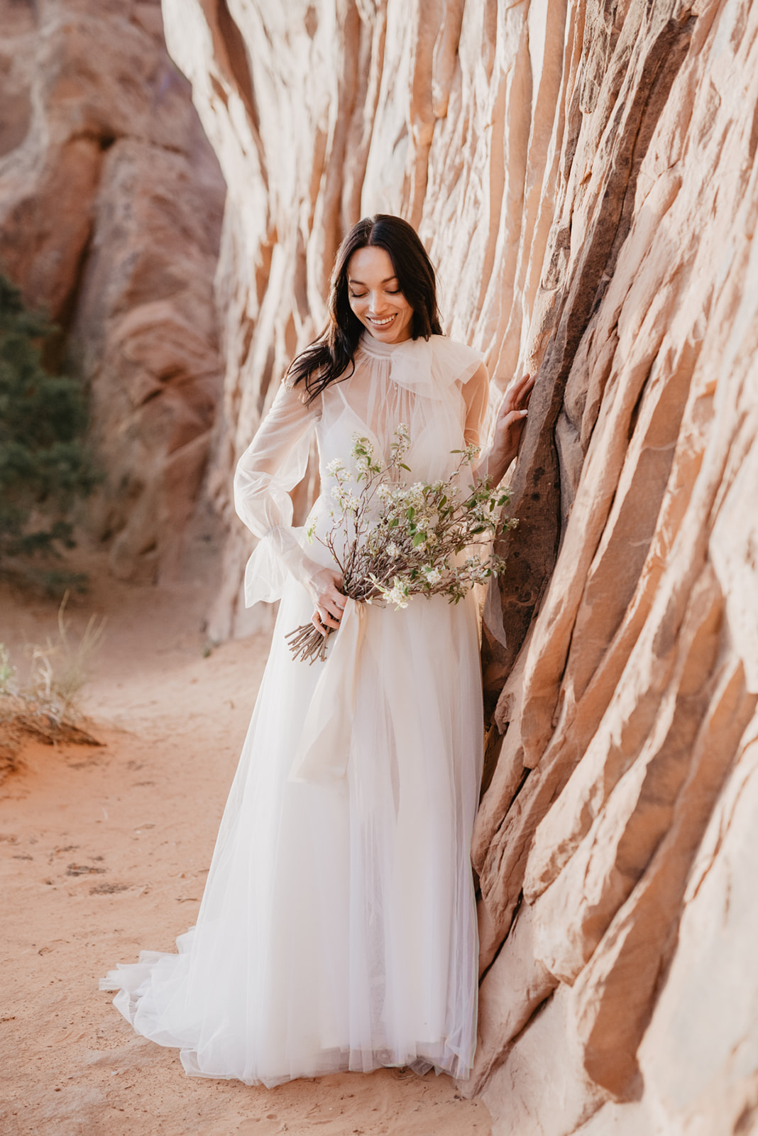 Utah Elopement Photographer photographs bride in a long sleeve wedding dress leaning against red rock at Arches National Park wedding