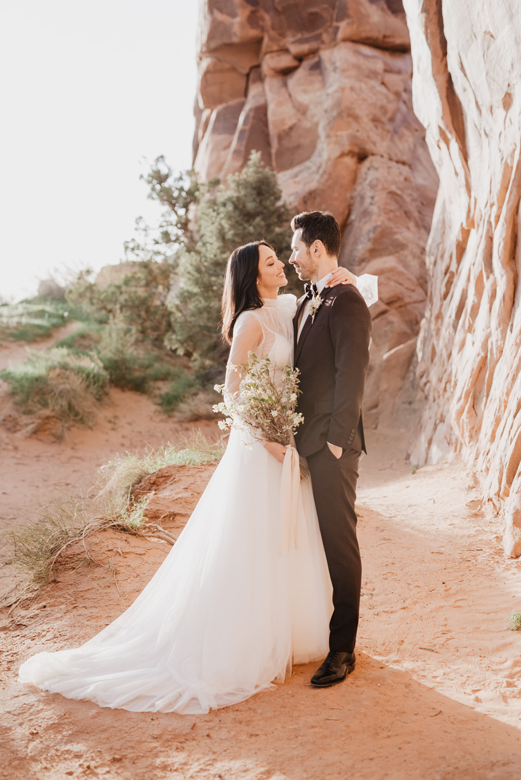 bride and groom embracing in red rocks of Southern Utah as the bride holds the grooms neck and smiles up at him while the sun sets in the distance captured by Utah elopement photographer