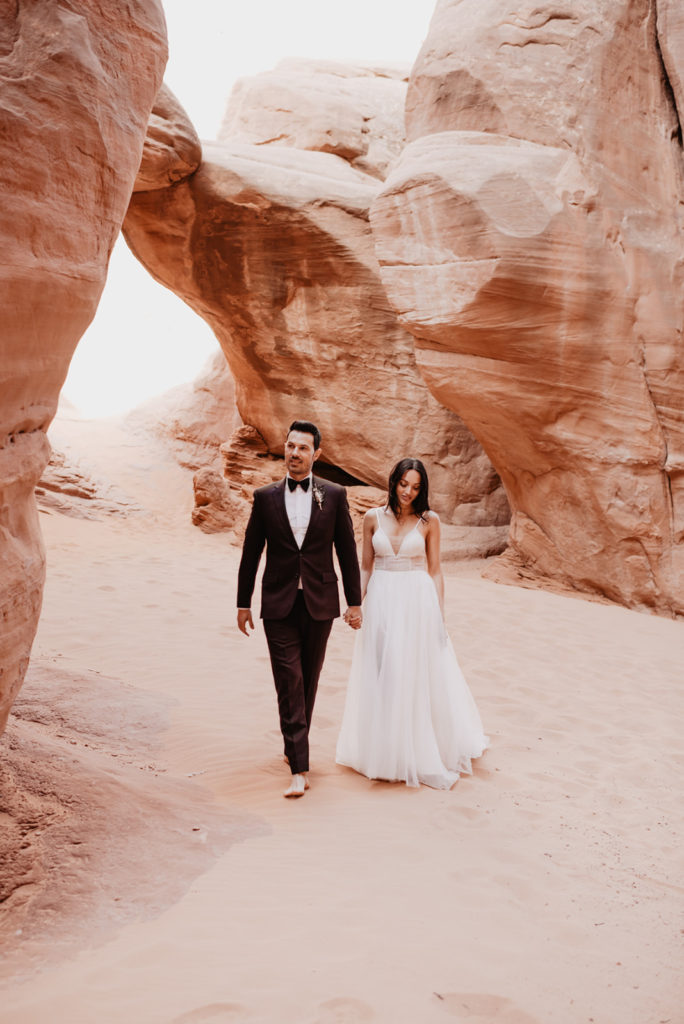 Utah elopement photographer captures bride and groom holding hands and walking in southern Utah's red rock for their Arches National Park wedding 