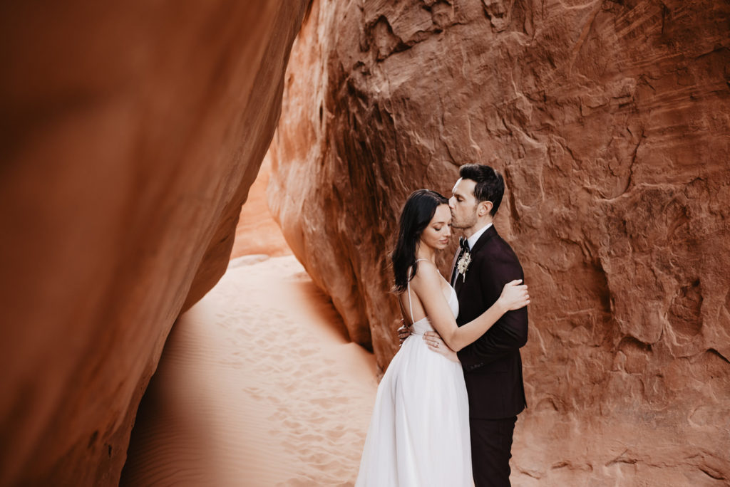 bride and groom embracing against red rock for their southern utah wedding with utah elopement photographer as the groom kisses his brides forehead while she leans into him 