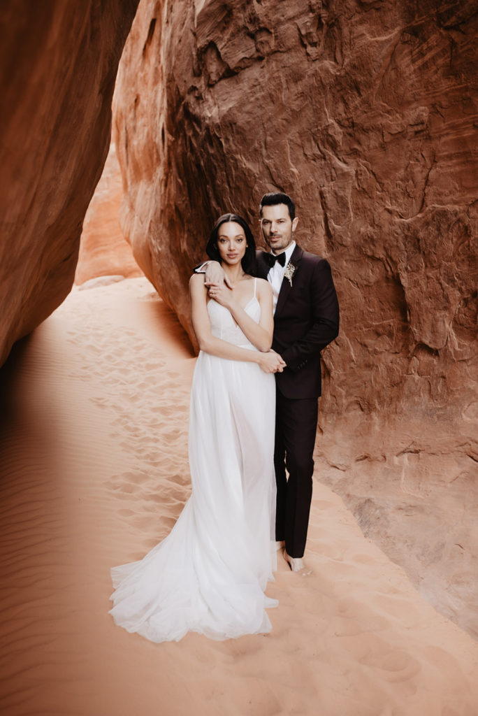 groom standing behind his bride while holdingher hands and smoldering with the sun shining through the caves captured by Utah Elopement photographers
