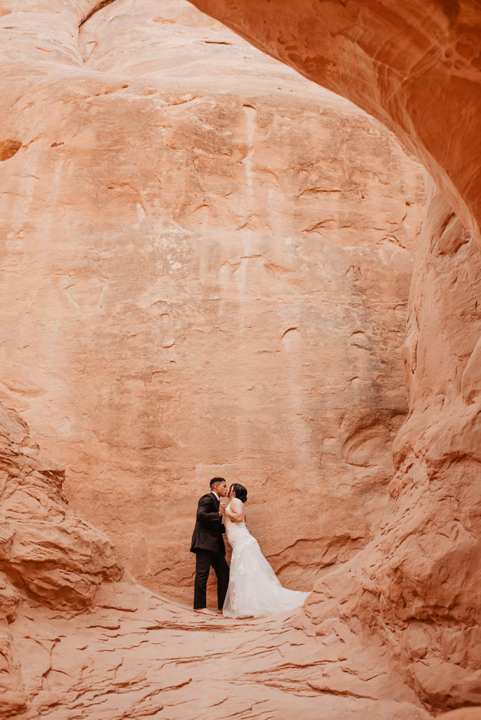 outdoor bridal in Arches National Park wedding photos with bride and groom in a red rock cave kissing captured by Utah elopement photographer