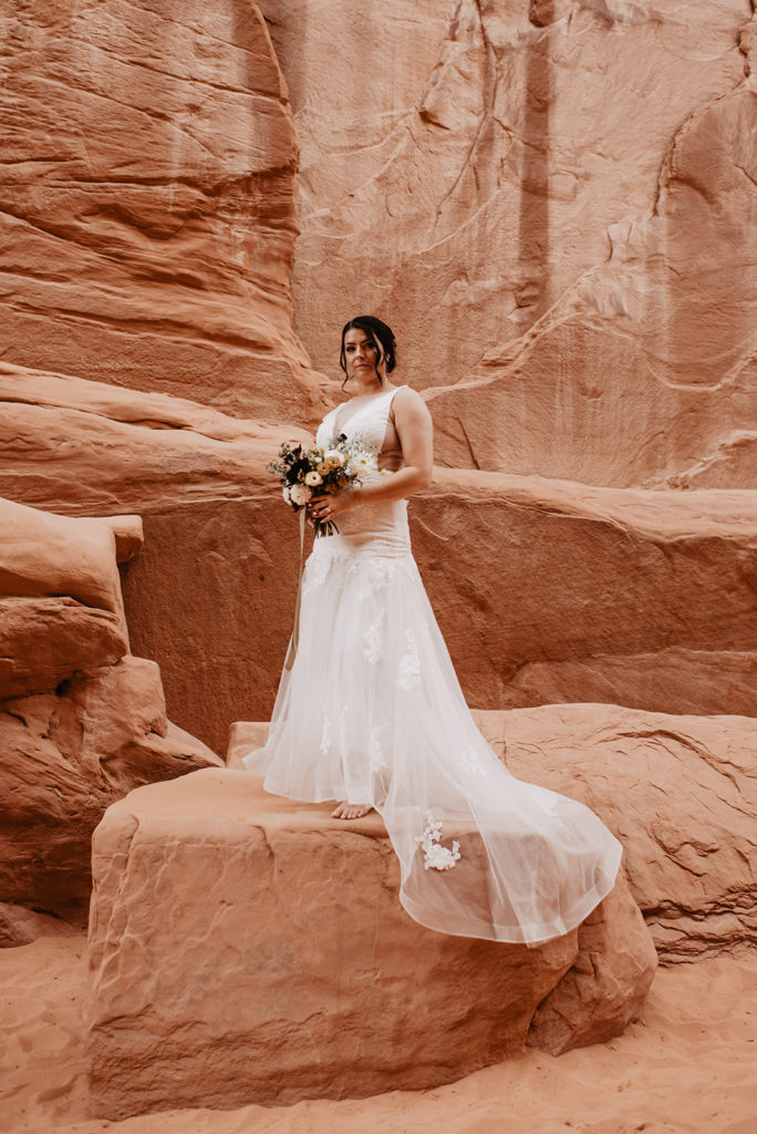 Arches National Park wedding with bride standing on a red rock while holding her floral wedding bouquet captured by Utah elopement photographer