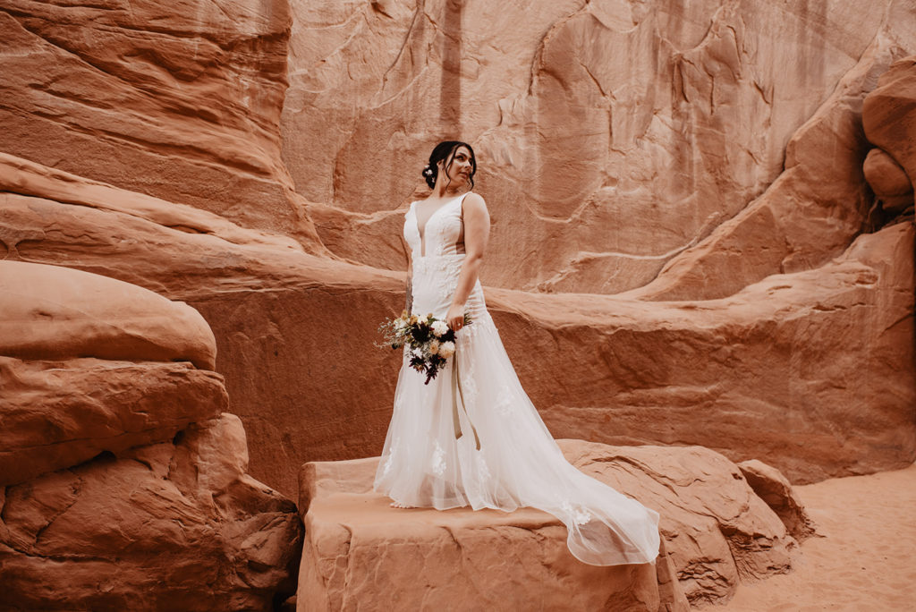 bride in a lace wedding dressing standing on a red rock in southern Utah while holding her floral bouquet in front on her down at her side and looking over her shoulder photographed by Utah elopement photographer 