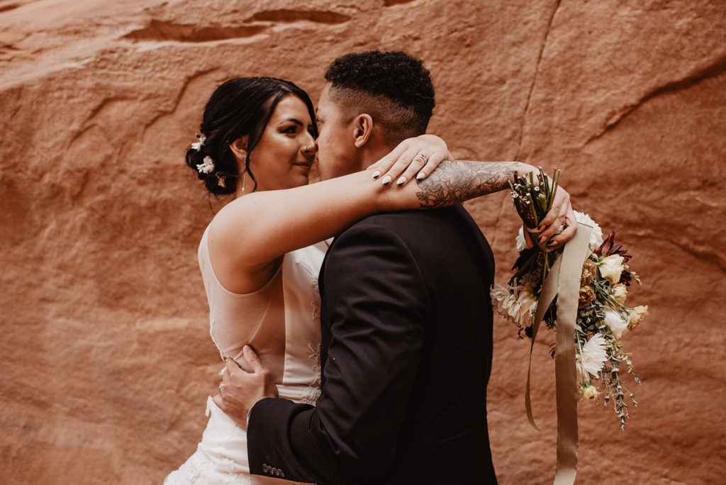 utah elopement photographer captures bridal portraits in Moab with bride and groom in a red rock cave as the bride throws her arms over the grooms shoulders and holds her bouquet to his back and he embraces her waist and leans in 
