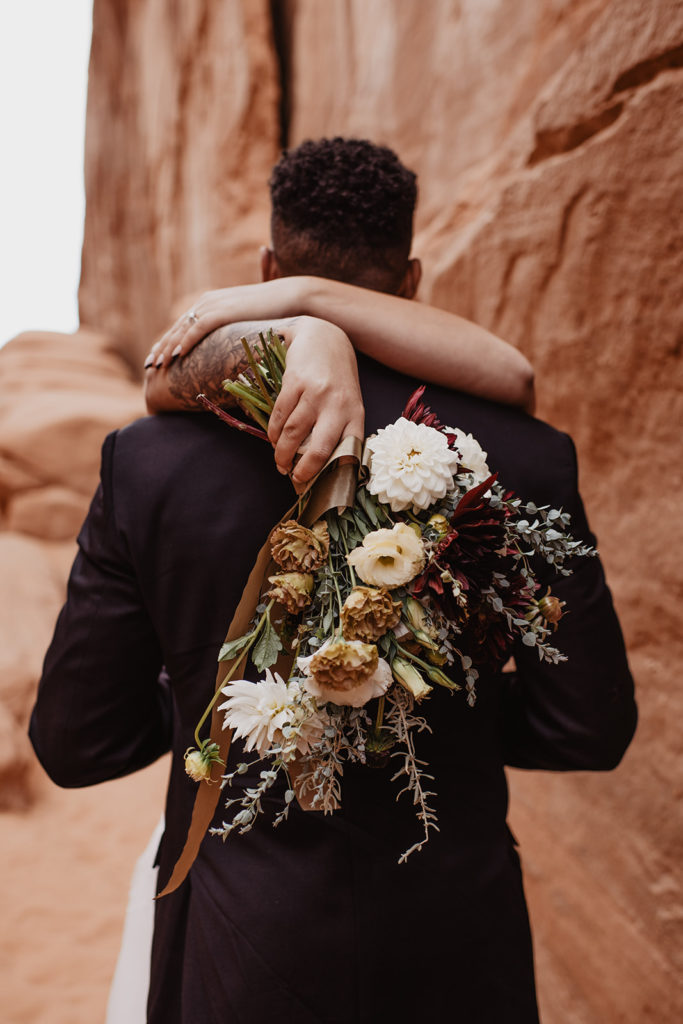 Southern Utah elopement photographer captures bride holds her arms around her grooms neck as her wedding bouquet hangs over his back for their Arches National Park wedding 