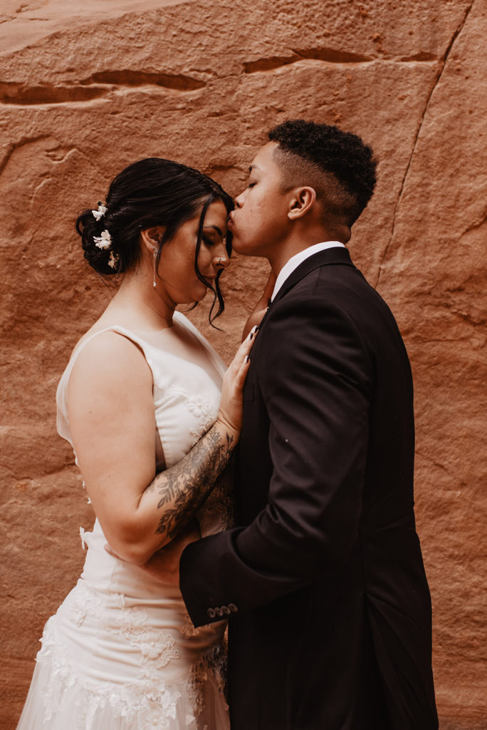 bridal portrait at red rock arches national park wedding with groom kissing his bride on the forehead while his bride leans into the grooms chest captured by Utah elopement photographer 