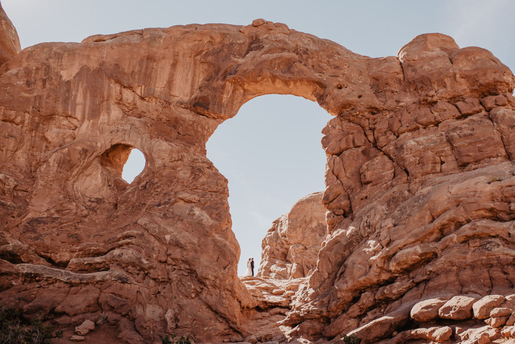 Utah elopement photographer captures bride adn groom embracing under a large arch at their Arches National Park wedding 