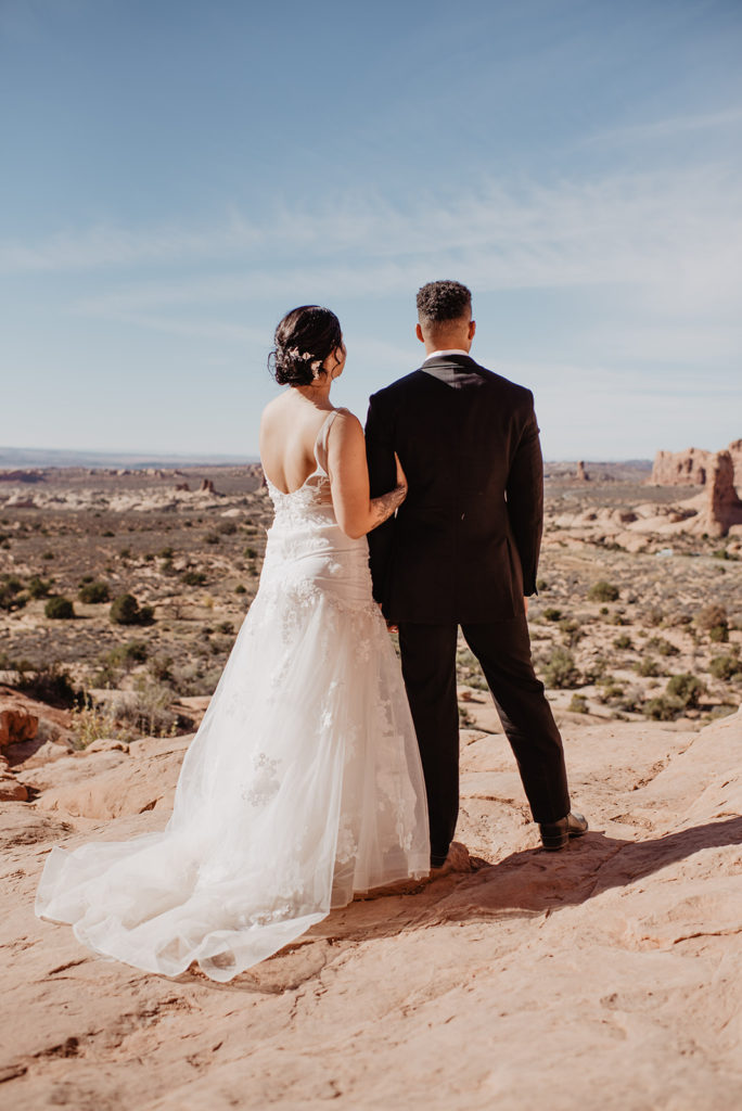 bridal portraits for Arches National Park wedding with bride and groom standing together on a red rock cliff while bride holds grooms arm and they look into the valley 