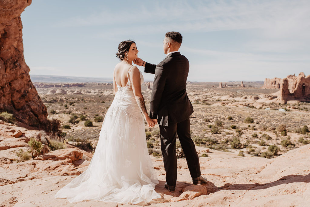 arches national park wedding pictures with utah elopement photographer capturing bride and groom hooding hands and standing on a mountain outlook in the red rocks while the groom reaches over with his other hand to caress his brides cheek 