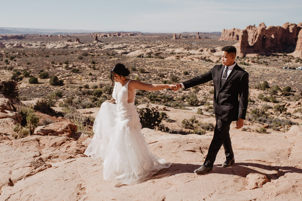 Utah elopement photographer captures bride and groom holding hands and walking over a cliff together with a view of Arches National Park 