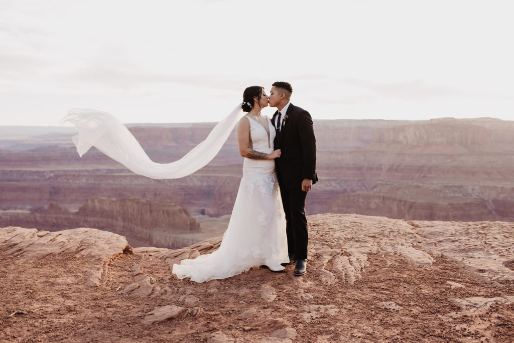 bridal photos for Arches National Park wedding with bride and groom kissing on a large red rock while the brides veil blows in the wind behind her at sunset with Utah elopement photographer