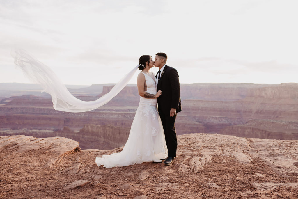 bride and groom standing at the top of a mountain at Arches National Park wedding while the sun sets in the distance and the brides veil flows in the wind captured by utah elopement photographer
