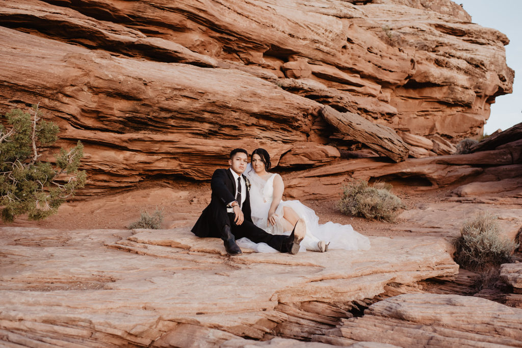 arches national park wedding photographer capture bride and groom sitting on red rock together in Moab in a canyon for their utah wedding pictures