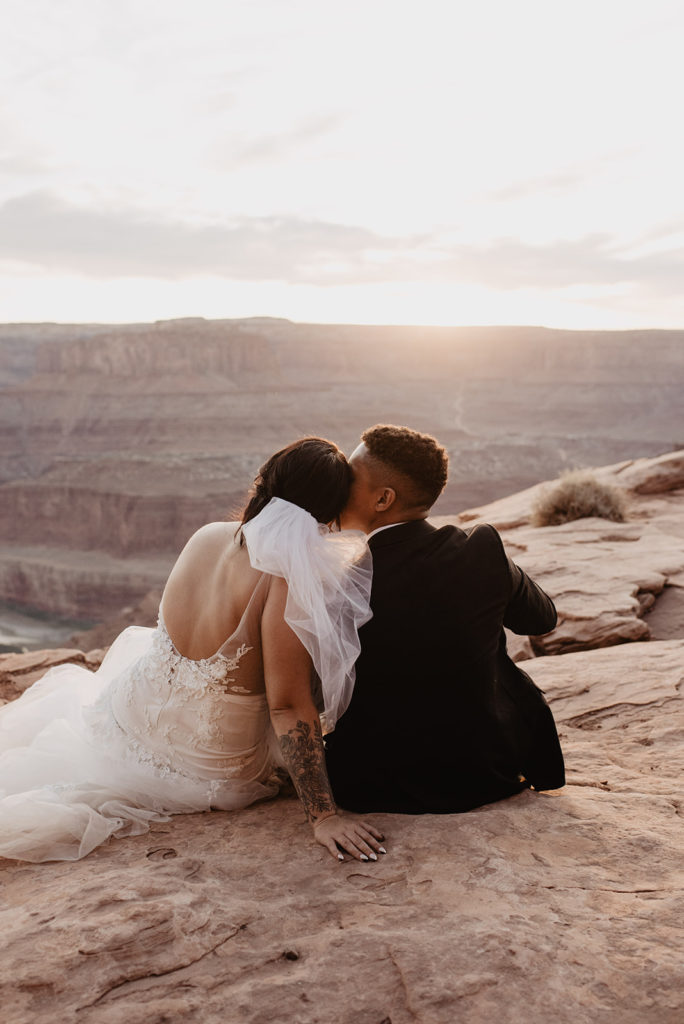 sunset wedding with bride and groom sitting together on a mountain top while the sun sets over the horizon as the groom leans over and kisses his bride for their Arches National Park wedding
