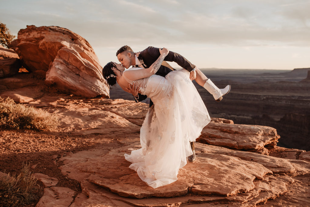 utah elopement photographer captures wedding portrait of groom dipping his bride backwards and holding her leg up as they kiss passionately as the sun shines while it sets 