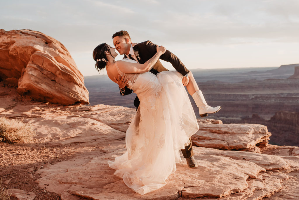 groom dips his bride backwards and leans in to kiss her as she holds his arm and the sun sets over them for their Utah Elopement 