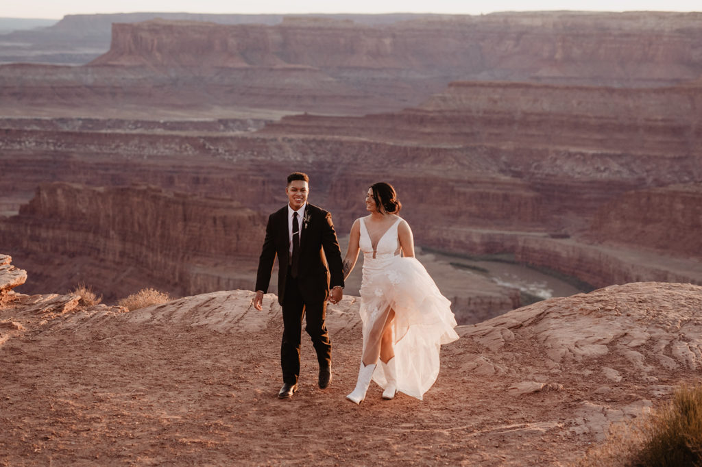 wedding portraits in Arches National Park with bride and groom holding hands and walking on the red rock cliffs together while the sunsets captured by Utah elopement photographer 