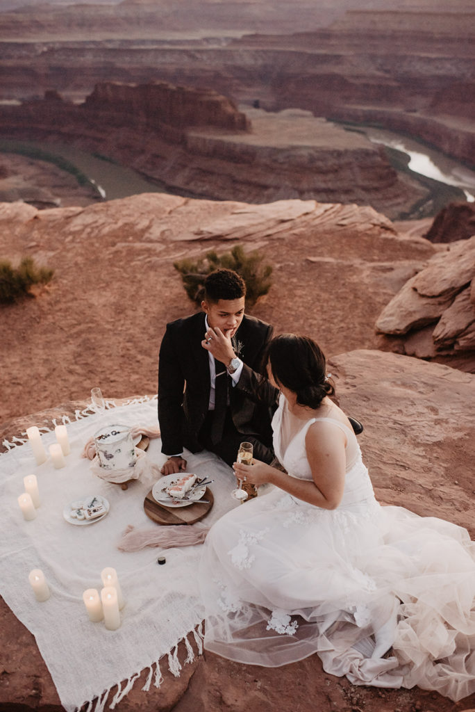 bride and groom having a picnic for their Arches National Park wedding as they sit on a white blanket together and eat cake captured by Utah elopement photographer