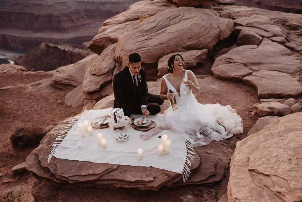 wedding picnic with bride and groom laughing as they eat wedding cake together on a white blanket on top of a mountain captured by utah elopement photographer 