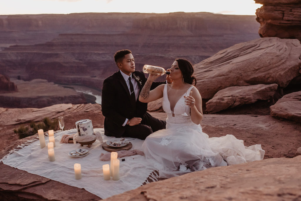 bride and groom drinking champagne together during a picnic on top of a mountain in southern Utah with candles surrounding them captured by Utah elopement photographer 