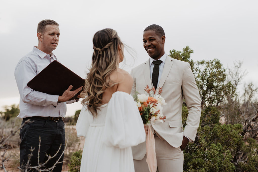 arches national park wedding photographer captures outdoor ceremony with bride and groom standing together and smiling as their officiant shares a few words