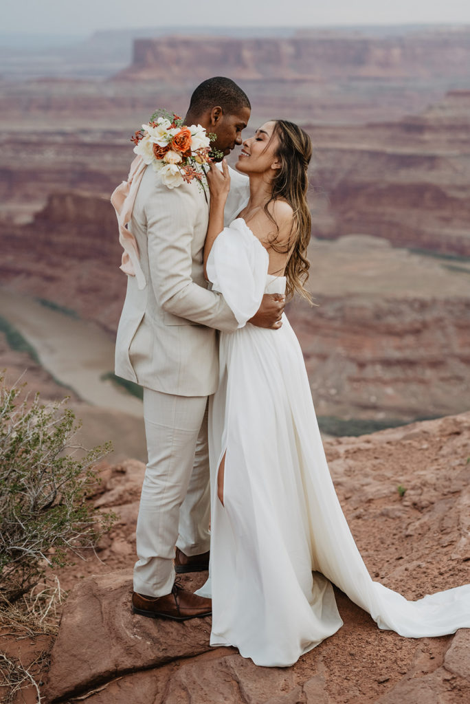 satin wedding dress on a beautiful bride as she embraces her groom and leans in to kiss his while she holds her floral bouquet over his shoulder captured by Utah elopement photographer 
