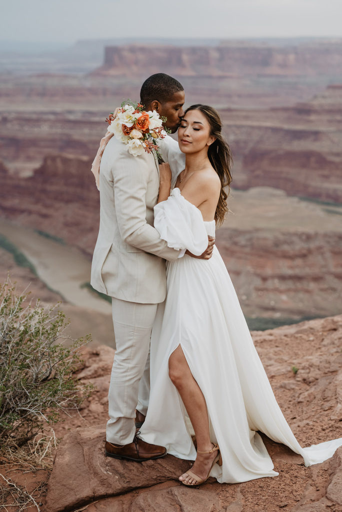 bride and groom embracing on a top of a mountain in Moab with groom in a tan suit holding his bride's waist as she looks over his shoulder as he kisses her temple with the canyon in the distance captured by Utah elopement photographer