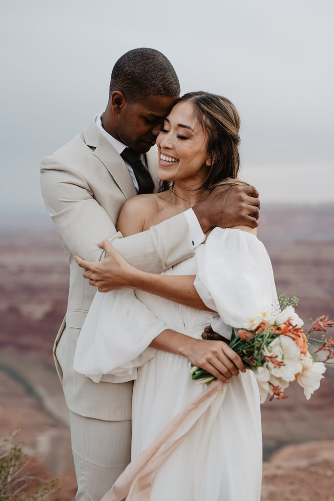 groom standing behind his bride with his arms around her shoulders and smiling down her face as he rests his head on her template while the bride holds her bouquet and reaches for her groom captured by utah elopement photographer 