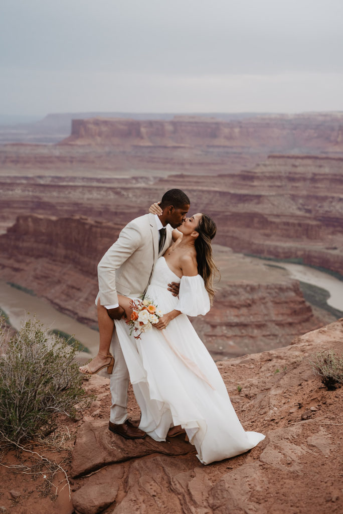 Utah elopement photographer captures bride and groom on top of a red rock mountain for their Arches National Park wedding with groom holding the brides leg up as the bride holds his neck 