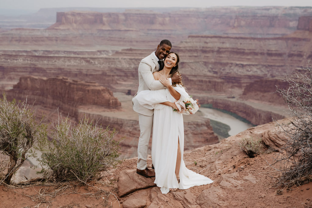 groom standing behind his bride and holding his arms around her waist and smiling as she leans into him with the canyon in Moab in the distance captured by Utah elopement photographer
