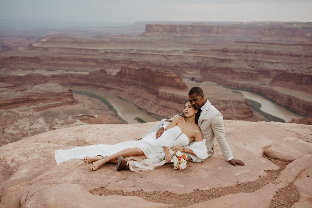 Utah Elopement Photographer captures groom sitting on a large red rock with his bride in his lap as she leans into him for romantic wedding pictures for Arches National Park wedding