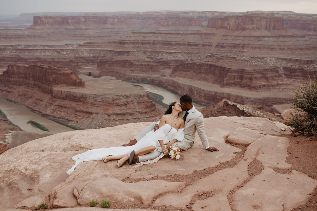 Utah elopement photographer captures bride and groom sitting together on red rock while the bride leans back into her groom and kisses him with a stunning canyon valley in the distance 