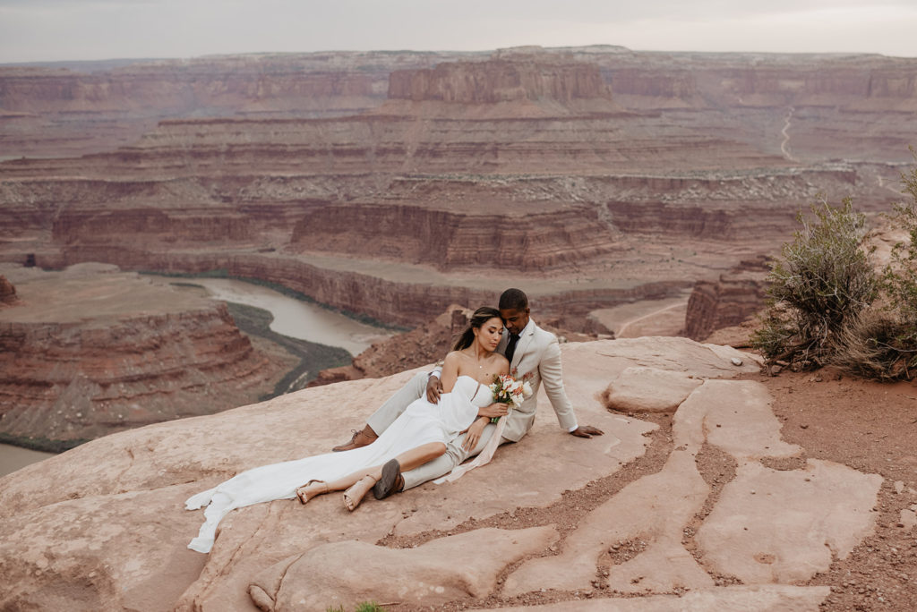 Arches National Park wedding with bride and groom sitting together on top of a cliff with a valley with a river in the distance photographed by Utah elopement photographer