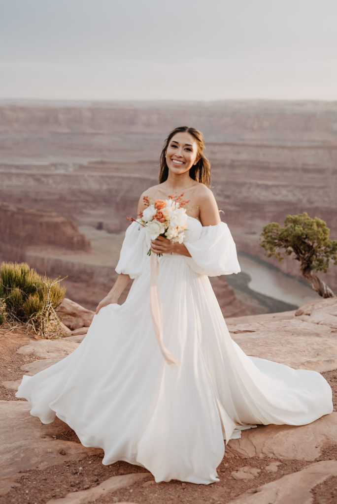 bride in a large tulle gown standing in red rock while holding her boho wedding bouquet as the wind blows her hair captured by Utah elopement photographer 