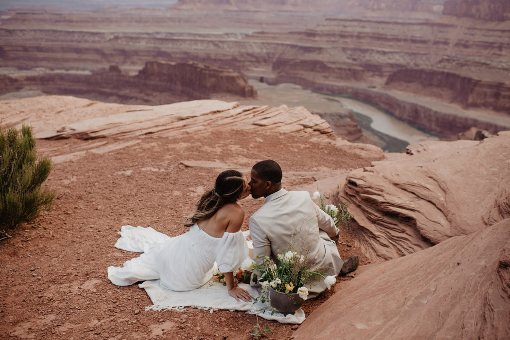 outdoor Arches National Park wedding with bride and groom sitting on top of a red rock mountain on a blanket and leaning into each other and kissing captured by Utah elopement Photographer