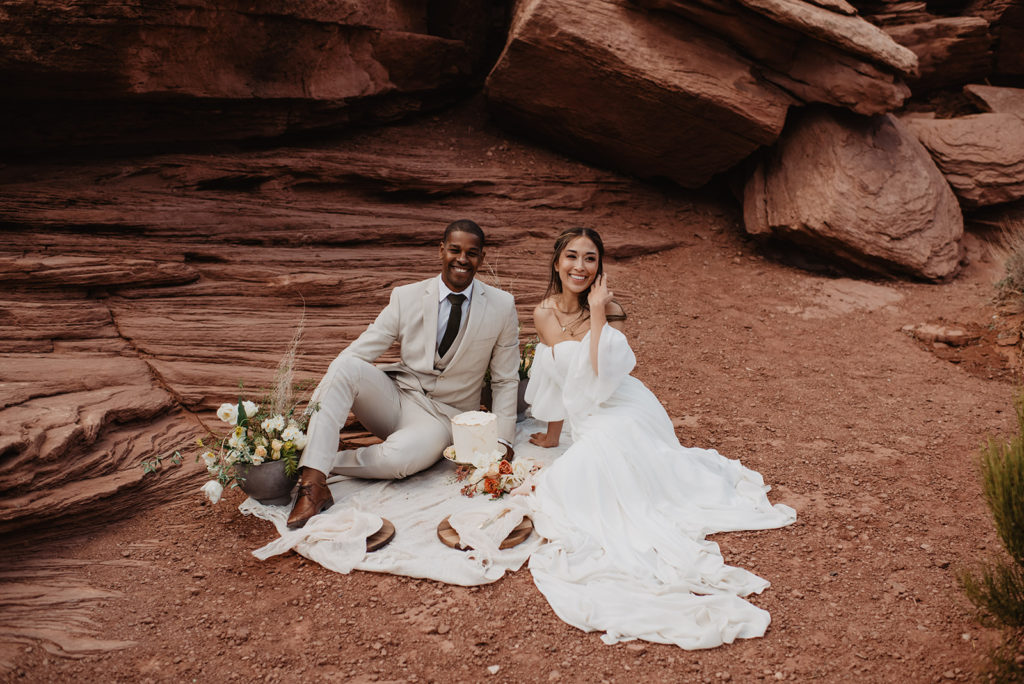 outdoor wedding with a picnic with bride and groom sitting on a blanket against red rock of Southern Utah with a small cake to celebrate captured by Utah elopement photographer