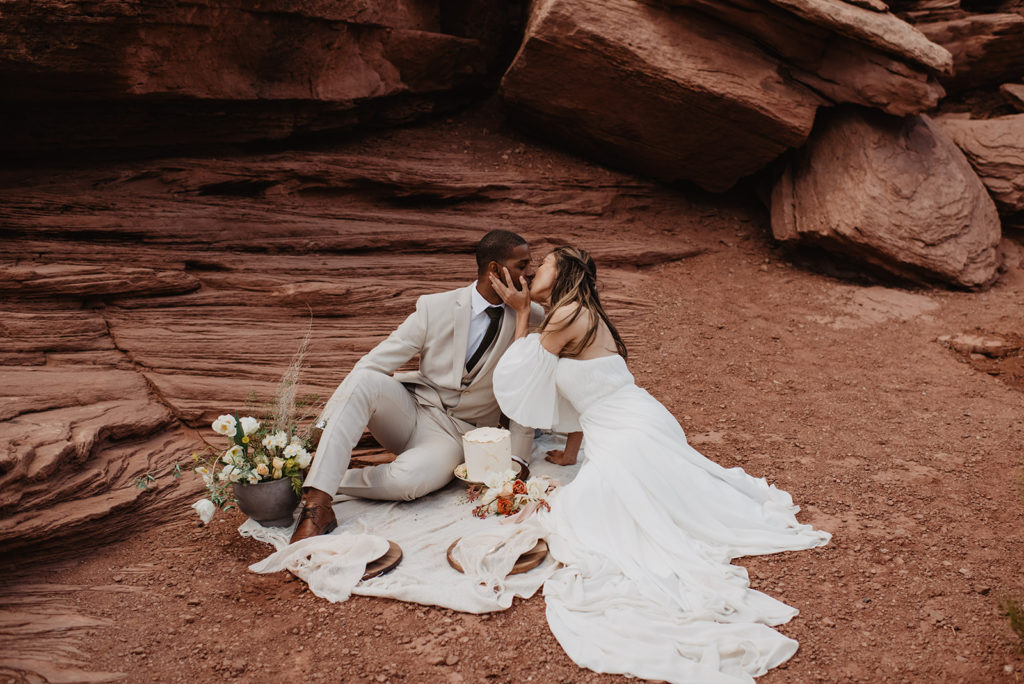 Utah elopement photographer captures wedding picnic in Arches National Park wedding with bride and groom kissing while sitting on a white blanket against the red rock with a small cake and florals