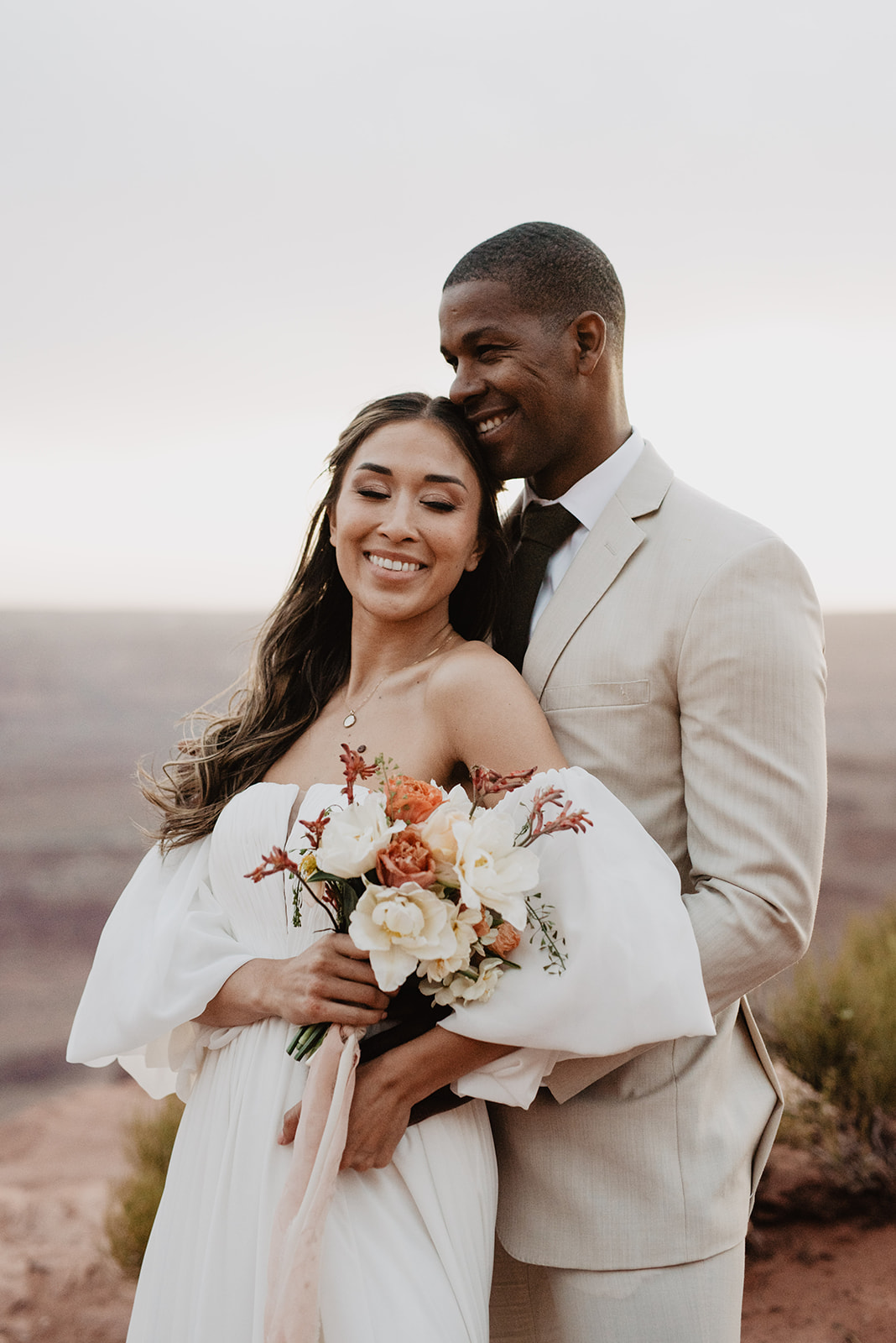 groom standing behind his bride as she leans back into him and smiles while she holds her floral wedding bouquet captured by Utah elopement photographer for Arches National Park wedding