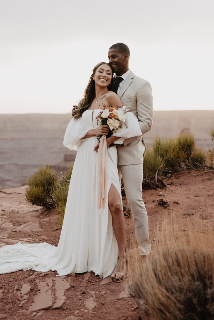 bride and groom embrace each other on a hill side in Arches National Park wedding with the groom standing behind the bride and holding her waist as she leans back into him and holds her floral bouquet to her shoulder 