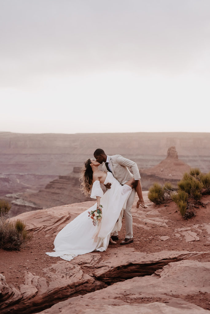 bride and groom on top of a canyon in Moab surrounded by red rocks with the groom dipping his bride back and holding her one leg and he kisses her passionately captured by utah elopement photographer 