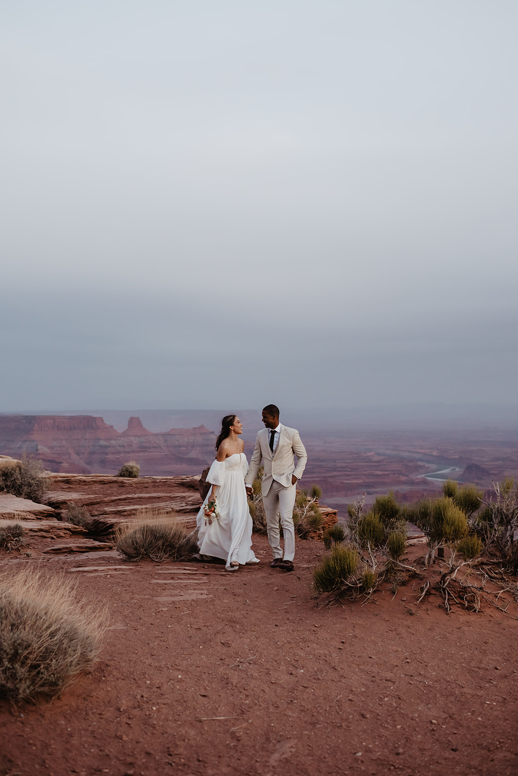 Utah elopement photographer captures bride and groom holding hands and walking at twilight on a cliff top with shrubs around them