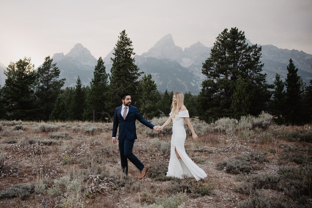 jackson hole photographers photographs Grand Teton wedding picture of groom holding his brides hand and leading her through a field with the Tetons in the distance