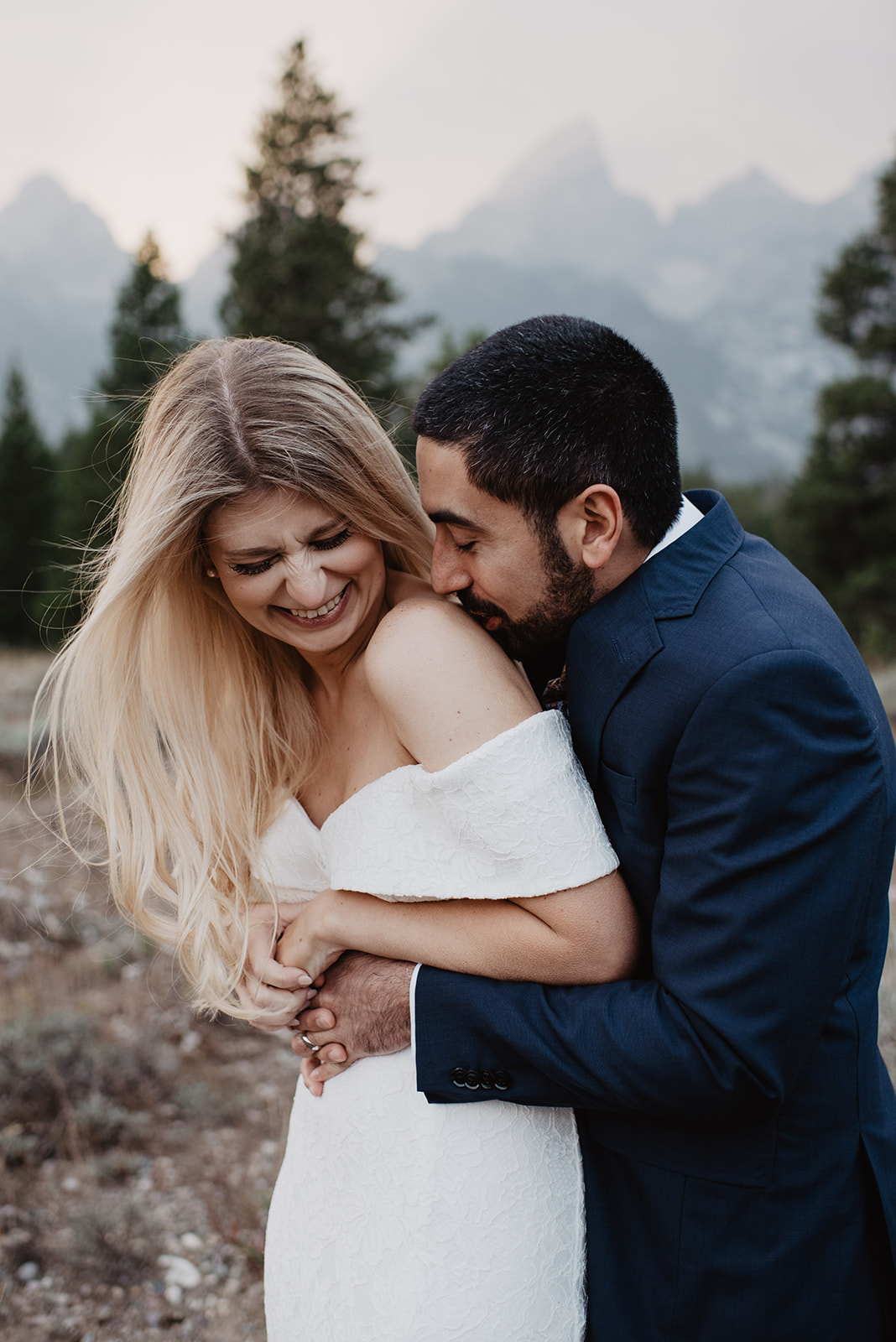 jackson hole photographers captures groom standing behind his bride and holding her around the waist and kissing her shoulder as she laughs