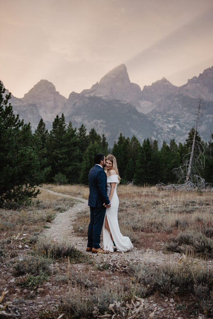 jackson hole photographer captures bride and groom embracing on a trail at teton glacier turnout with a dusty pink sky above the mountains 