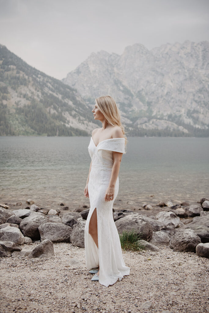 Jackson Hole photographers captures bride in a satin gown standing on a lake shore with large rocks surrounding her as she looking out across the lake for her outdoor bridal photos