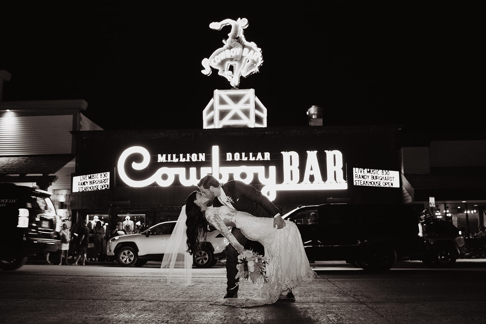 Jackson Hole photographers captures groom dipping his bride backwards and kissing her passionately in the middle of the road with the Cowboy Bar of Jackson Hole behind them with the lights turned on after dark