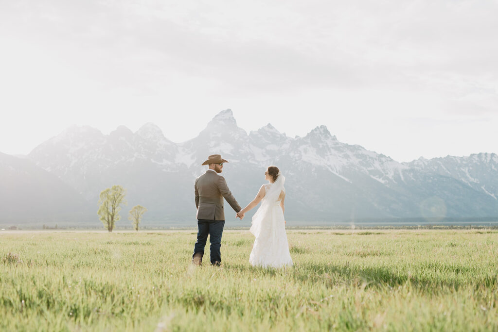 Wyoming elopement photos with bride and groom holding hands as they walk towards the grand tetons in the distance in an open field with the sun beginning to set for their Grand Teton wedding photos