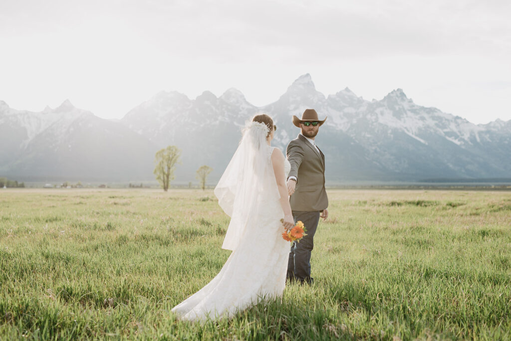 jackson hole photographers photographs bride and groom holding hands with the groom leading the bride through a field as he looks back to his bride with the tetons in the distance