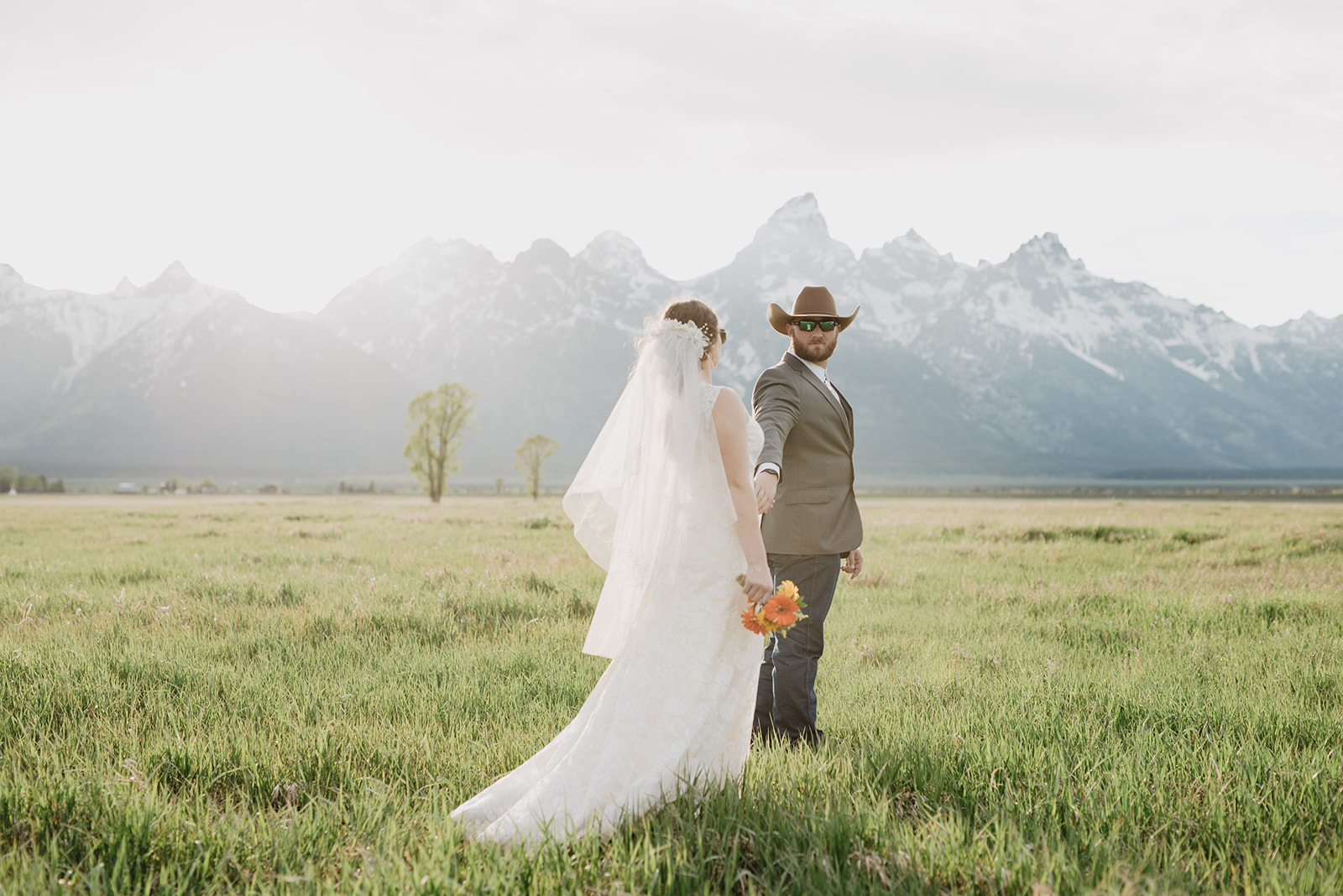 jackson hole photographers photographs bride and groom holding hands with the groom leading the bride through a field as he looks back to his bride with the tetons in the distance